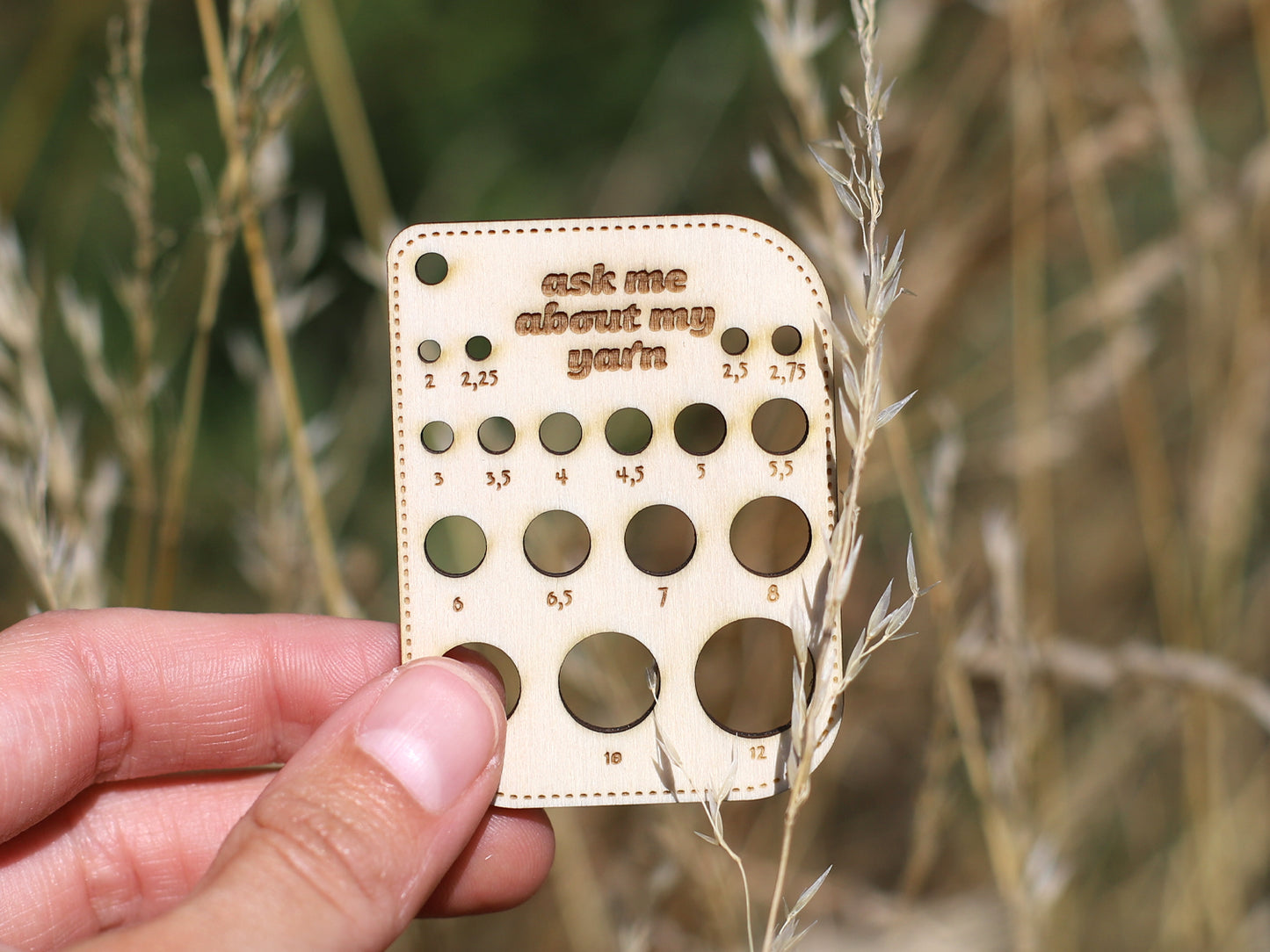wooden knitting needle size gauge held in a hand with grass background, features holes for measuring needle sizes.