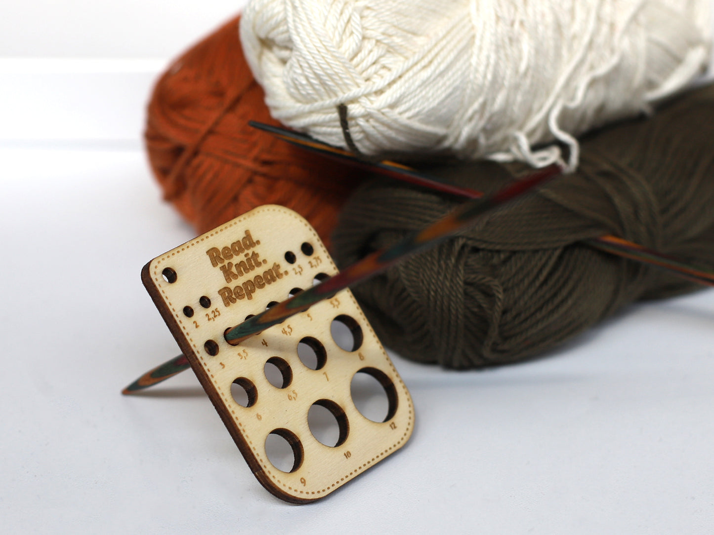 Kleines Nadelmaß für Stricknadeln mit Holzdesign und bunten Wollknäueln im Hintergrund.