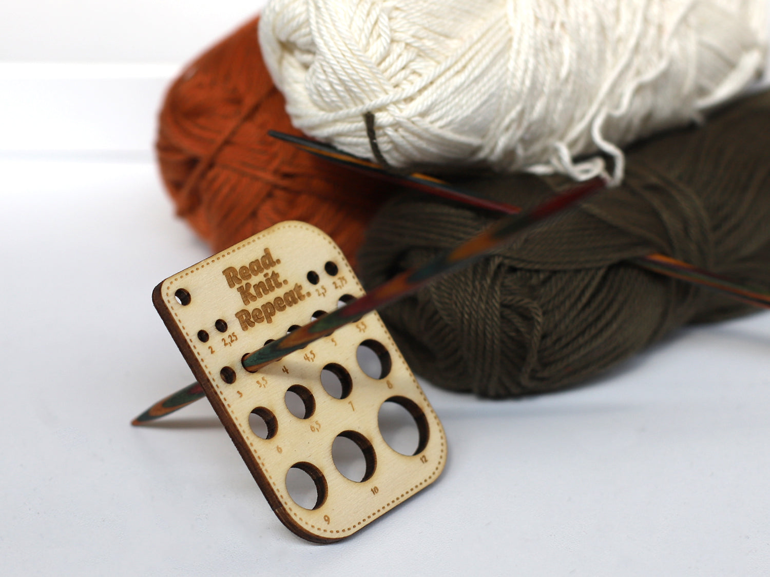 Kleines Nadelmaß für Stricknadeln mit Holzdesign und bunten Wollknäueln im Hintergrund.